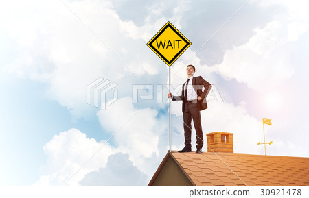 Young businessman on house brick roof holding yellow signboard. Mixed media 30921478