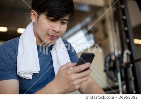 Young Asian man using mobile phone in the gym Young Asian man using mobile phone in the gym 30958382