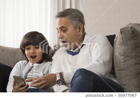 Happy grandfather and grandson using mobile phone sitting together on sofa Happy grandfather and grandson using mobile phone sitting together on sofa 30977067