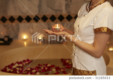 A staff is holding a candle in a bathroom with bathtub, flowers, candles. 31285013