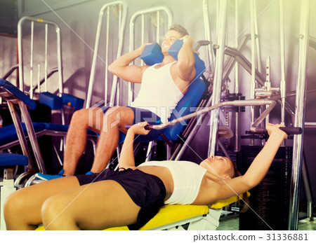 Young vigorous woman lying on back on bench and lifting weights in shoulder press machinery Young vigorous woman lying on back on bench and lifting weights in shoulder press machinery 31336881