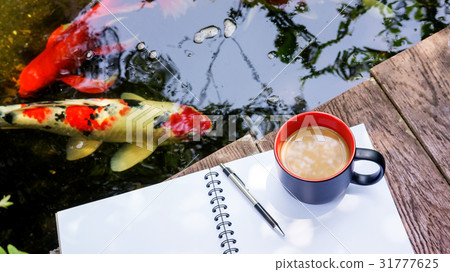 hot coffee, book, and pen place near a fancy carp pond. 31777625
