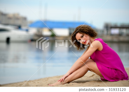 Young woman in a red dress ans curly hair sit on the water coast 31933120