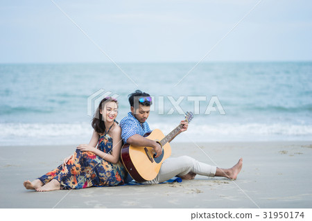 A man is sitting close his girlfriend and playing guitar on the beach 31950174