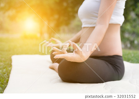 asian pregnant woman practicing yoga while sitting in lotus position on green grass in public park. meditating on maternity. concept of healthy lifestyle and relaxation. 32031552