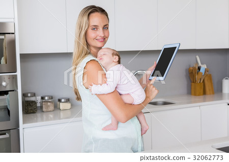 Mother using digital tablet while carrying her baby in kitchen 32062773