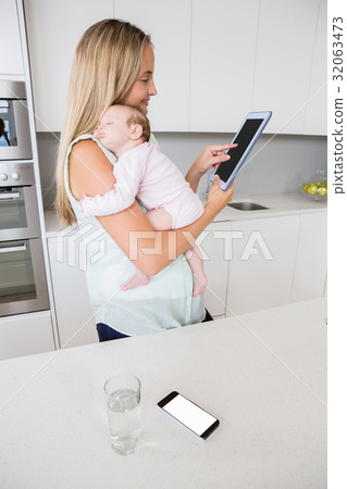 Mother using digital tablet while carrying her baby in kitchen 32063473