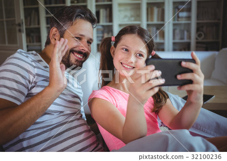 Smiling father and daughter looking at mobile phone in living room 32100236