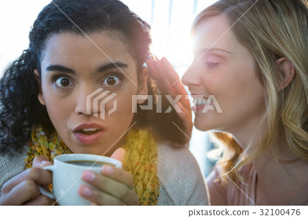 Woman whispering a secret into her friends ear while having coffee 32100476