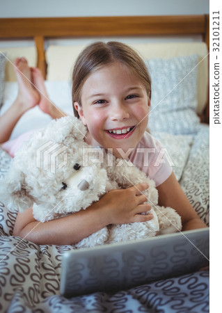 Happy girl lying on bed with a teddy bear and using digital tablet Happy girl lying on bed with a teddy bear and using digital tablet 32101211