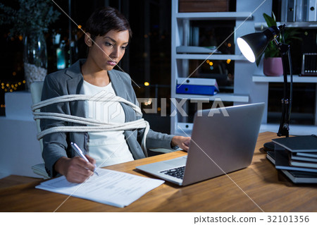 Businesswoman tied with rope while working on laptop at her desk 32101356