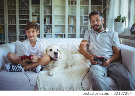 Father and son sitting on sofa with pet dog and playing video games 32107294