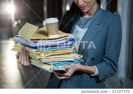 Businesswoman carrying stack of file folders while using mobile phone Businesswoman carrying stack of file folders while using mobile phone 32107370