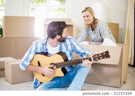Man playing a guitar while woman unpackaging cardboard boxes in background Man playing a guitar while woman unpackaging cardboard boxes in background 32255399