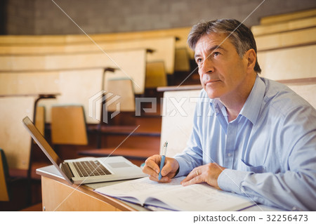 Thoughtful professor writing in book at desk 32256473