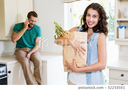 Woman holding groceries while man talking on mobile phone in kitchen Woman holding groceries while man talking on mobile phone in kitchen 32258501