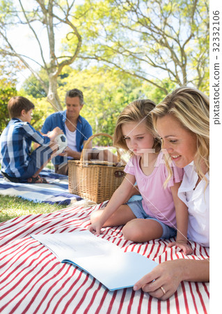 Mother and daughter reading book while father and son playing with football in park 32332106