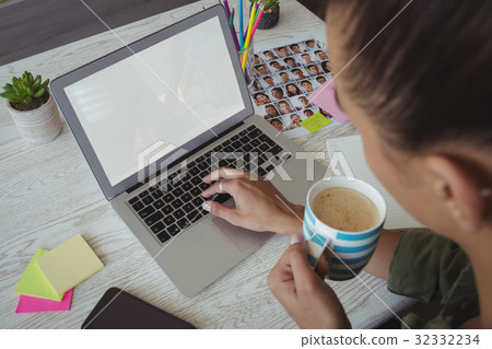 Female photo editor holding coffee cup while using laptop in office Female photo editor holding coffee cup while using laptop in office 32332234