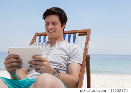 Man sitting on sunlounger and using digital tablet on the beach Man sitting on sunlounger and using digital tablet on the beach 32332374