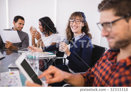 Smiling businesswoman with drinking glass sitting by colleagues Smiling businesswoman with drinking glass sitting by colleagues 32333318