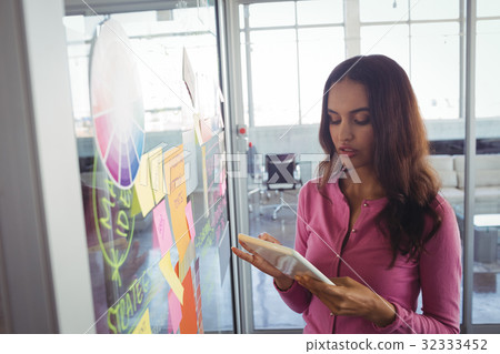 Female designer holding digital tablet by adhesive notes on glass in office Female designer holding digital tablet by adhesive notes on glass in office 32333452