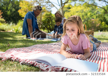 Girl lying on blanket and reading book while family sitting in background Girl lying on blanket and reading book while family sitting in background 32333584