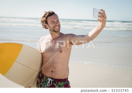 Smiling shirtless man taking selfie with surfboard at beach 32333745