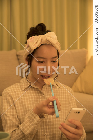Woman eating potato chips with tongs at night while looking at a smartphone 33005979