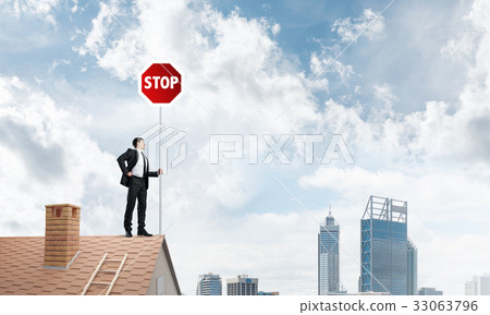 Caucasian businessman on brick house roof showing stop road sign. Mixed media Caucasian businessman on brick house roof showing stop road sign. Mixed media 33063796