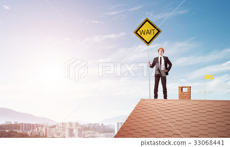 Young engineer on house brick roof holding yellow signboard and looking at city. Mixed media 33068441