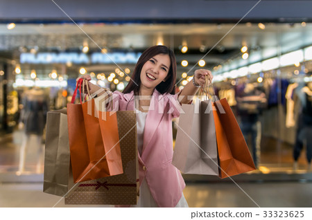 Happy young asian woman shopping in happy feeling and holding the product paper bag over the Abstract blurred photo of clothing store in a shopping mall, shopping concept Happy young asian woman shopping in happy feeling and holding the product paper bag over the Abstract blurred photo of clothing store in a shopping mall, shopping concept 33323625