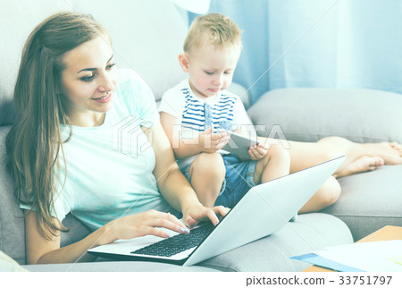 Satisfied mother is productively working behind laptop while child playing on tablet Satisfied mother is productively working behind laptop while child playing on tablet 33751797