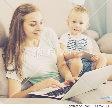 Smiling mother is productively working behind laptop while child playing on tablet Smiling mother is productively working behind laptop while child playing on tablet 33752121