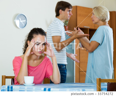 Sad young woman watching husband and mother having fight Sad young woman watching husband and mother having fight 33754272