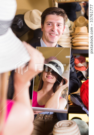 assistant holding mirror and showing woman his reflection in hats store 33761777