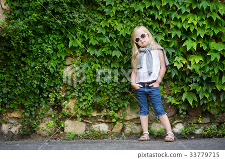 Funny little girl wearing sunglasses posing by bindweed wall on warm and sunny summer day Funny little girl wearing sunglasses posing by bindweed wall on warm and sunny summer day 33779715