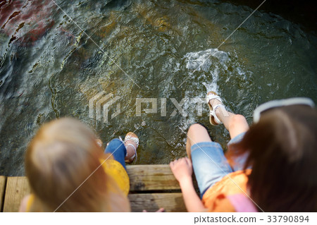 Two cute little girls sitting on a wooden platform by the river or lake 33790894