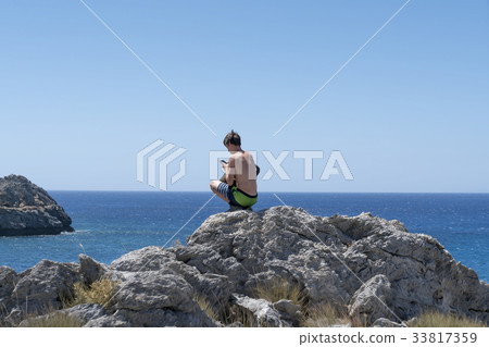 young man crouching on a rock with mobile phone 33817359