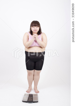 Fat young woman standing in front of a weight scale with praying in front of white background 33849699