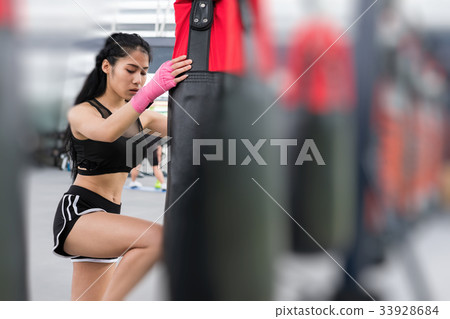 young woman execute exercise in fitness center. female athlete hitting and punching sandbag with rowing machine in boxing gym. sporty girl working out in health club. martial arts, muay thai concept young woman execute exercise in fitness center. female athlete hitting and punching sandbag with rowing machine in boxing gym. sporty girl working out in health club. martial arts, muay thai concept 33928684
