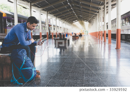 asian man with backpack sitting on platform at train station. backpacker or traveler with headphone look at mobile phone while waiting for train. journey, trip, travel concept 33929592