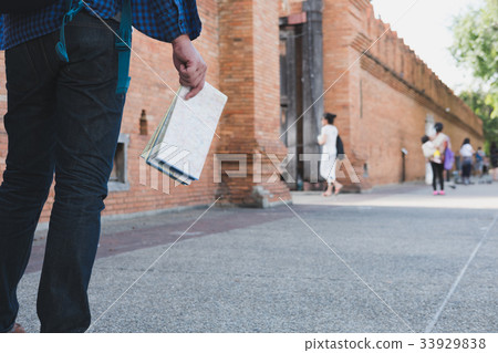 young asian man wearing blue shirt and jeans with map and backpack standing near old brick wall young asian man wearing blue shirt and jeans with map and backpack standing near old brick wall 33929838
