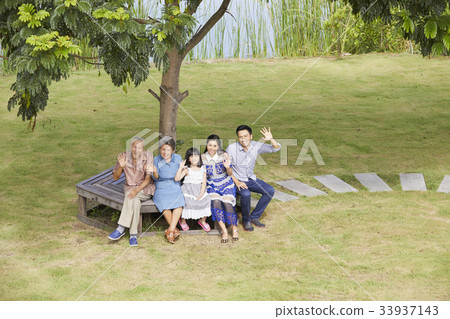 a family is sitting under a tree, waving their hands and looking at camera 33937143