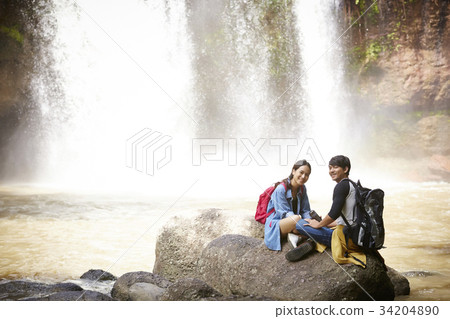 A happy couple is sitting on the rock near the waterfall. 34204890