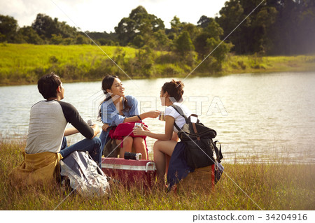 Three friends are sitting and drinking coffee on the riverbank. 34204916