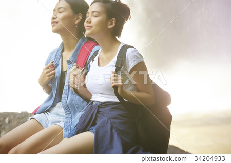 Two young woman are relaxing while sitting near the waterfall 34204933