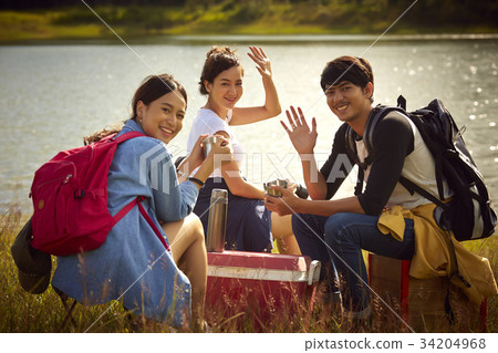 A group of friends is sitting, smiling and waving hand on the riverside 34204968