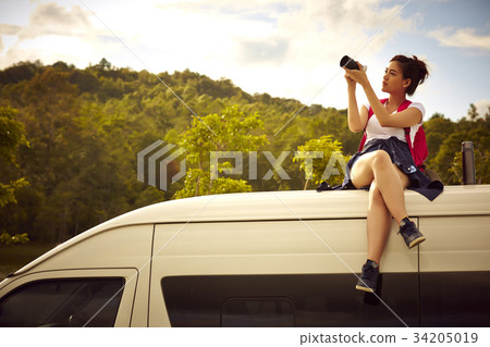 a female traveler is sitting on a top of a car, taking pictures with digital camera 34205019