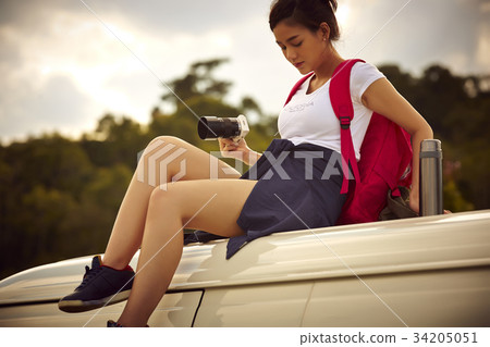a young female traveler is sitting on a top of a car and holing digital camera 34205051