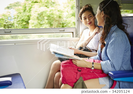 Two Asian girls are sitting and looking at each other on a train. 34205124